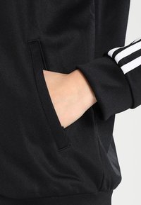 Sudadera atlética negra con un bolsillo lateral y puños acanalados, fabricada con un tejido suave y texturizado, y con franjas de acento blancas en la manga.