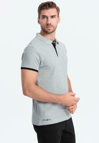 Šedé polo tričko s černým límečkem a akcenty na rukávech, s jemným logem na spodním lemu. Vyrobeno z hladkého, prodyšného materiálu.