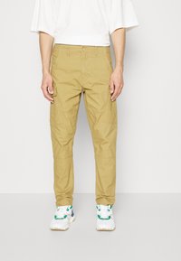 Beige cargo kalhoty s tapered střihem, se bočními kapsami a knoflíkovým zapínáním, v kombinaci s bílými teniskami se zelenými akcenty.