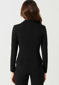 Blazer noir ajusté à manches longues, avec revers crantés et taille cintrée. Tissu lisse et design minimaliste sans dispositifs visibles.