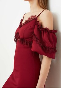 Femme portant une robe bordeaux à épaules dénudées avec bordure en dentelle et manches bouffantes, se tenant contre un mur blanc.