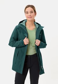 Giacca impermeabile verde scuro con cappuccio, dotata di tasche con cerniera, interno a contrasto verde chiaro, abbinata a una camicia verde chiaro.
