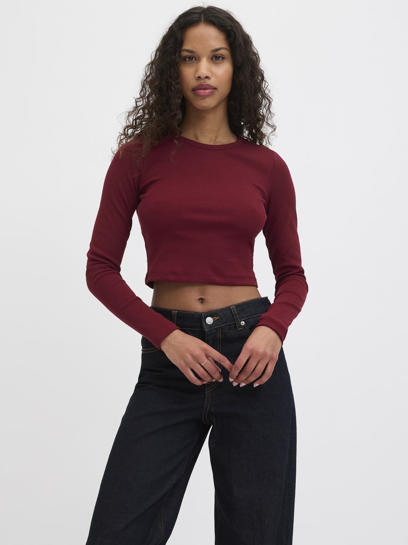 Langermet, cropped topp i burgunder ribbestrikket stoff, parret med mørk denim høyt liv bukser med knapper og sømdetaljer.