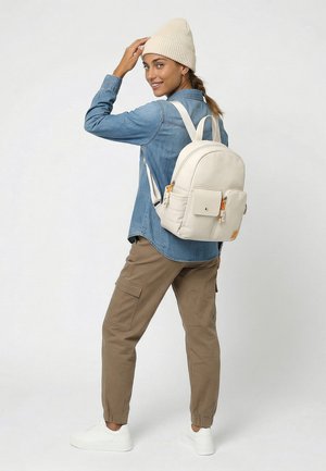 Nuori nainen beige pipo päässään, sininen farkkupaita ja ruskeat cargo-housut, kantaa kermaista reppua, hymyilee ja katsoo olkapäänsä yli taaksepäin.