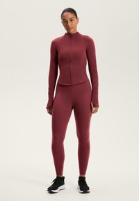 Burgundy sportos szett, amely cipzáros, hosszú ujjú felsőből és hozzá illő leggingsből áll. Rugalmas anyagból készült, sima felülettel, fekete sportcipőkkel viselve.