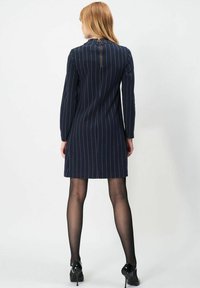 Marineblå strikket kjole med lodrette hvide striber, lange ærmer og høj halsudskæring. Lynlåsdetalje på ryggen, knælængde, parret med sorte hæle.