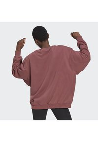 Person med kort hår som höjer båda armarna, iklädd en överdimensionerad sweatshirt i dammrosa och svarta byxor, sedd bakifrån mot en enfärgad bakgrund.