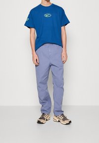 Man med en blå T-shirt med en gul logotyp, ljuslila byxor och beige- och svarta sneakers, ståendes mot en enkel ljus bakgrund.