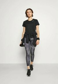 Sort atletisk t-shirt, mønstrede grå leggings, sorte sneakers. Modellen holder en vandflaske og har en sort hoodie bundet om livet.