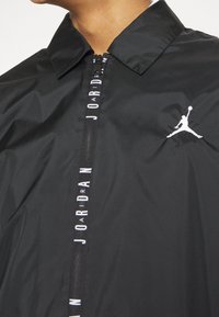 Černá bunda se zipem vpředu, kterou zdobí límec. Bílé logo "JORDAN" podél zipu a bílé logo Jumpman na hrudi.