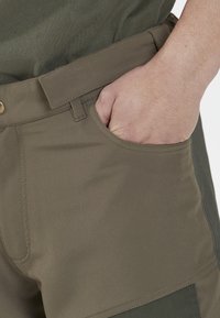 Oikea käsi liukuu khaki-housujen etutaskuun, joissa on samaa kangasta oleva vyö ja oliivinvihreä paita tukahtuneena.