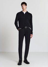 Schwarzer gerippter Strickpullover mit einem Viertel-Reißverschluss-Kragen, kombiniert mit schlank geschnittenen schwarzen Hosen und mattschwarzen Stiefeletten. Durchgehend glatte Textur.