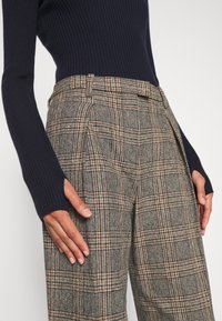 Femme portant un pantalon à carreaux marron taille haute et un pull côtelé bleu marine foncé ajusté à manches longues, mains reposant sur les côtés.
