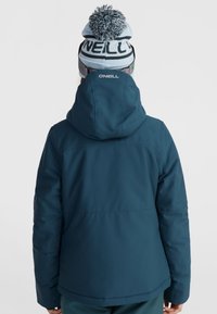 Veste isolée turquoise avec capuche, présentant une texture lisse et un design à l'arrière. Bonnet en tricot noir et blanc avec un pompon.