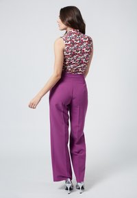 Ärmelloses blumiges Top in Rot, Grün und Weiß, kombiniert mit hoch taillierten lila Hosen; das Model steht mit dem Rücken zur Kamera, silberne Absätze sind sichtbar.