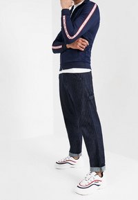 Navy tracksuit jacket with red and white stripe detail, paired with dark pinstriped pants and white sneakers with red accents.