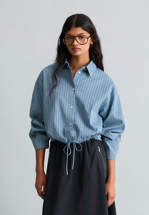 Young woman with long dark hair wearing large glasses, a blue striped button-up blouse with drawstring waist, and a black skirt with zipper pockets.