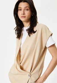 Top avvolgente senza maniche beige con una finitura testurizzata, caratterizzato da un dettaglio con bottone di lato e indossato sopra una t-shirt bianca.