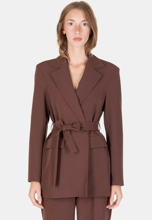 Donna che indossa un blazer marrone con cintura e rever, abbinato a pantaloni coordinati, in piedi davanti a uno sfondo chiaro uniforme.