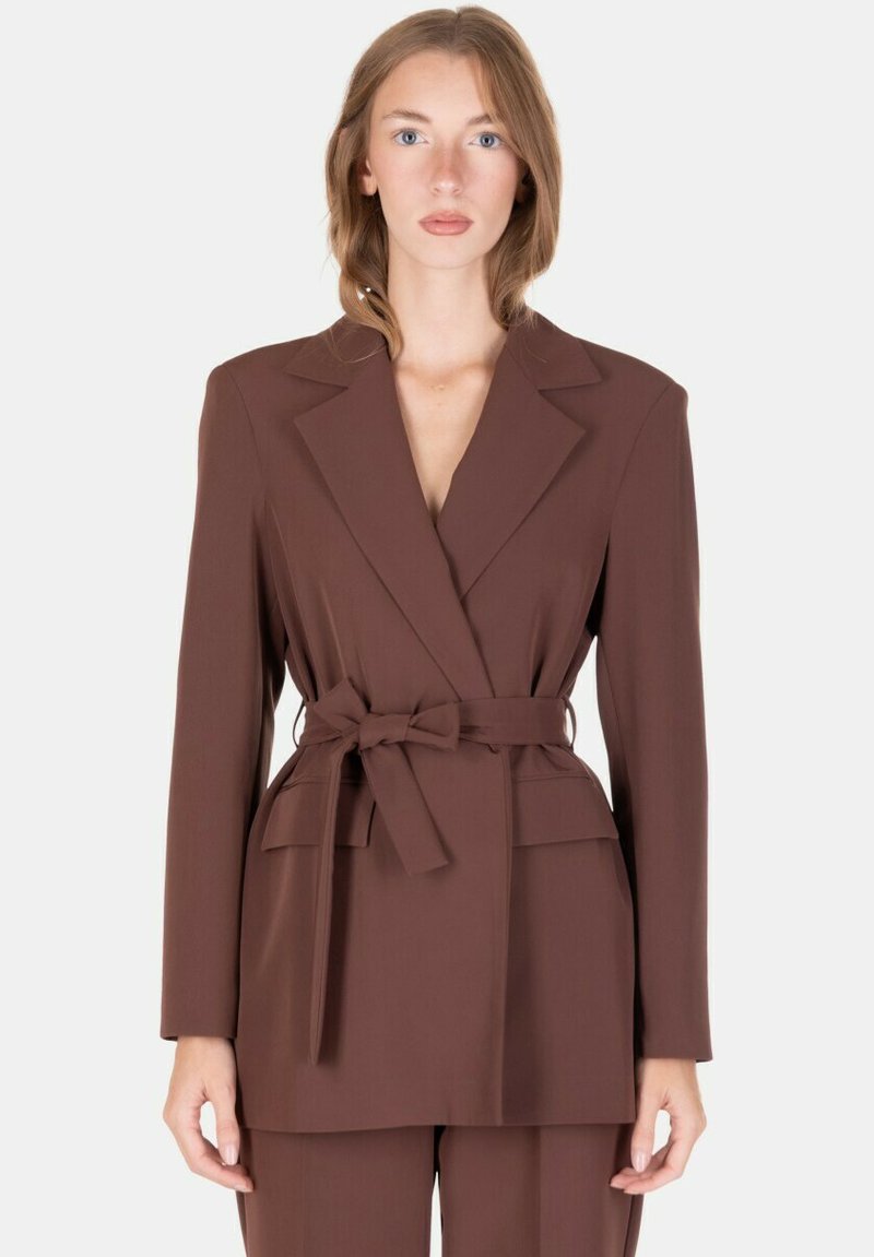 Donna che indossa un blazer marrone con cintura e rever, abbinato a pantaloni coordinati, in piedi davanti a uno sfondo chiaro uniforme.