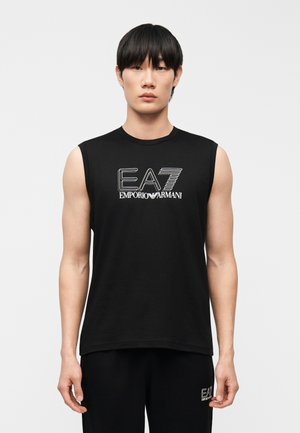 Modèle masculin portant un débardeur noir EA7 Emporio Armani et un pantalon noir assorti, sur un fond clair uni.