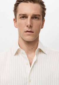 Camisa blanca con rayas verticales, cuello abotonado, tela suave y ligero brillo. El modelo mira directamente a la cámara.