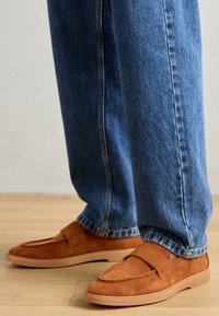 Braune Wildlederloafer mit einer flachen, hellbraunen Sohle, ausgestattet mit einem gestickten Design und einem Stoffriemen über der Vorderseite, kombiniert mit blauen Jeans.