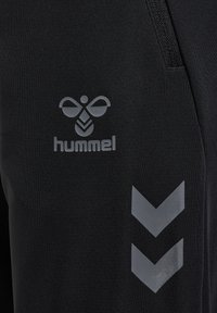Svarta träningsbyxor i stretchmaterial med en grå "hummel"-logotyp och tre chevron-accenter i matta nyanser.