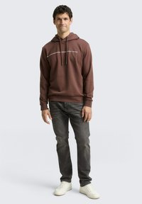 Bruine hoodie met trekkoorden en witte tekst, gecombineerd met donkere jeans en witte sneakers. Eenvoudig ontwerp en casual pasvorm.