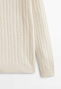 Manche et bas d'un pull en maille torsadée couleur crème avec poignets côtelés et motifs verticaux texturés sur fond blanc.