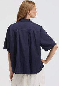 Navy blue short-sleeve shirt with a relaxed fit, featuring a back yoke and pleats for added texture. Lightweight fabric.
