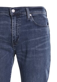 Jean en denim bleu foncé à coupe slim, design cinq poches, coutures contrastantes et fermeture par bouton métallique à la taille.