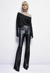 Schwarzes Schulterfreies Langarmoberteil kombiniert mit hochtaillierten schwarzen Leder-Hosen mit weitem Bein, verziert mit metallischen Beschlägen und Ösen.