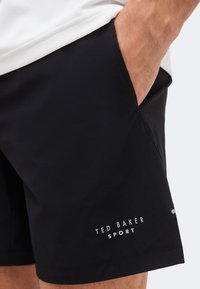 Κοντινό πλάνο ενός ατόμου που φοράει μαύρο σορτς Ted Baker Sport με το χέρι στην πλαϊνή τσέπη, σε απλό φόντο.