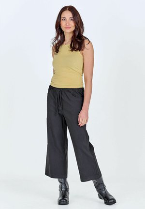 Jonge vrouw met lang bruin haar, gekleed in een mouwloze gele top, wijde zwarte broek en zwarte enkellaarzen, staande tegen een effen achtergrond.