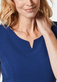 Marineblaues, strukturiertes Kleid mit V-Ausschnitt, das die Details des Stoffes zeigt und eine schmale Passform an den Schultern hat. Haut und Hand sind teilweise sichtbar.