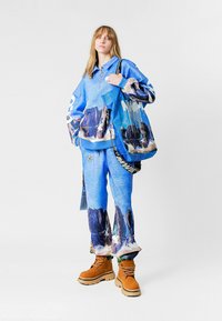 Sudadera azul con capucha y cremallera, con estampado gráfico de montañas. Pantalones azules a juego con el mismo estampado. Botas marrones con suelas gruesas y cordones.