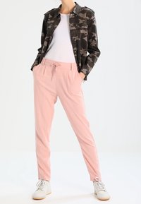 Kvinna som bär en kamouflagejacka, vit skjorta, rosa dragsko-byxor och vita sneakers, stående med händerna i fickorna.