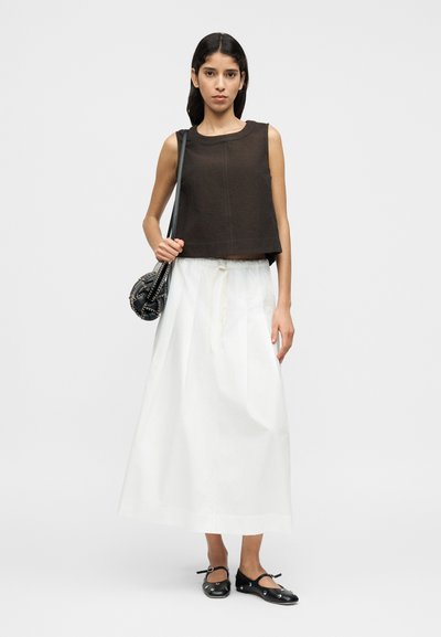 Καφέ κοντό αμάνικο τοπ με στρογγυλή λαιμόκοψη, σε συνδυασμό με λευκή μακριά φούστα. Το μοντέλο κρατά μια μικρή μαύρη τσάντα με μεταλλικές λεπτομέρειες.