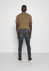 Brun kortärmad skjorta i kombination med mörkblå tajta jeans. Jeansens bakfickor har orange sömmar och tyget har slitna detaljer.