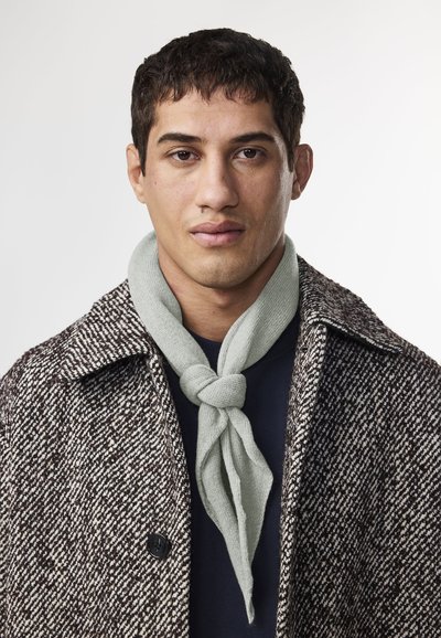 Cappotto a bardot marrone con bottoni neri, indossato sopra una camicia blu navy. Una sciarpa verde chiaro è annodata attorno al collo.