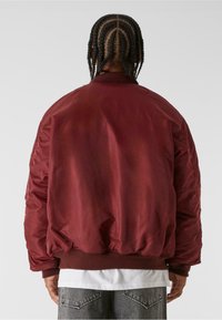 Bordová bomber bunda s hladkou texturou, žebrovanými manžetami a spodním lemem, má uvolněný střih a vycpané ramena. Zadní pohled ukazuje bez log.