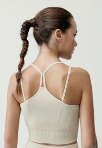 Beige sportovní podprsenka s nastavitelnými tenkými ramínky. Má ribbovanou texturu, široký spodní lem a minimalistický design s jemným brandingem na zadní straně.