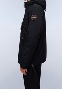 Sort jakke med en tekstureret overflade, der har en justerbar hætte, frontlommer og et rundt logo-patch på venstre arm.