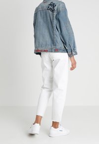 Osoba oblečená v modré denimové bundě s textem, bílých zkrácených džínách a bílých teniskách Reebok, stojící proti jednobarevnému pozadí.