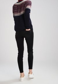 Marineblauer Pullover mit mehrfarbigem Fair-Isle-Muster an den Schultern, kombiniert mit schlanken schwarzen Jeans und weißen Sneakers mit goldenen Akzenten.