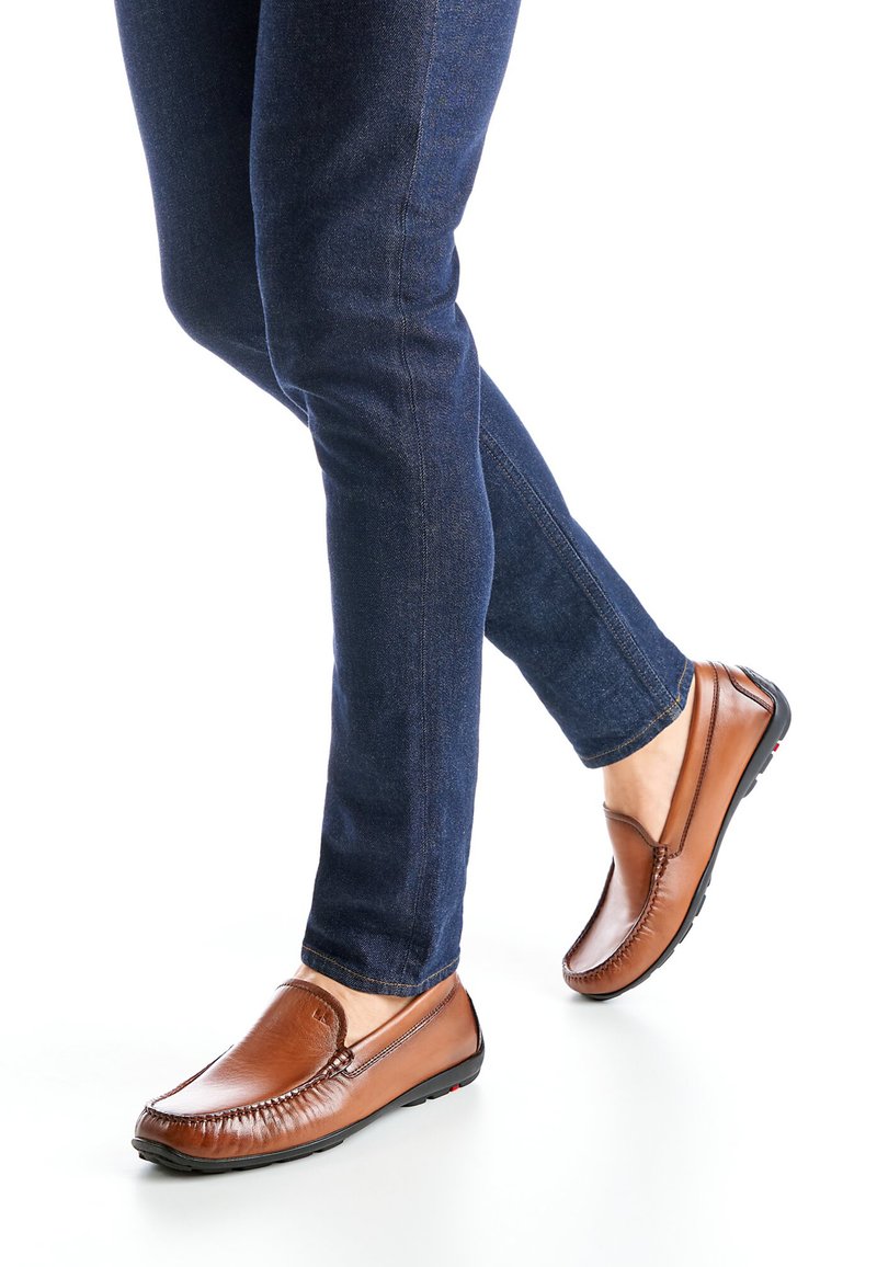 Braune Leder-Slipper mit Mokassinnaht, kombiniert mit dunkelblauen, eng geschnittenen Jeans, auf weißem Hintergrund.