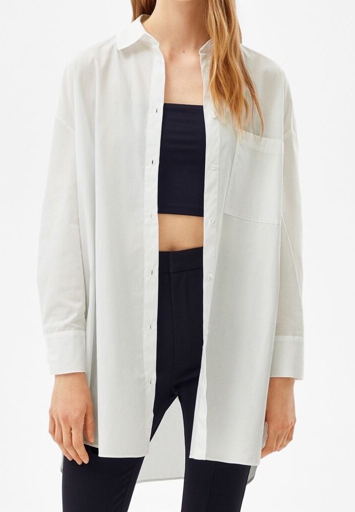 Femme portant une chemise blanche oversize à boutons par-dessus un crop top noir et un pantalon noir taille haute, debout devant un fond blanc.