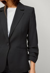 Zwarte op maat gemaakte blazer met gelapte revers, enkele knoopsluiting en opgerolde manchetten. Gladde stof met twee voorzakken.