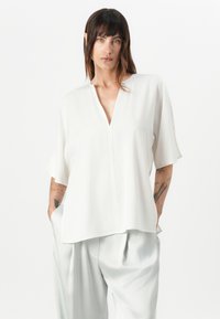 White short-sleeve blouse with a V-neckline, featuring a relaxed fit and soft, smooth texture. Paired with light gray, loose-fitting trousers.